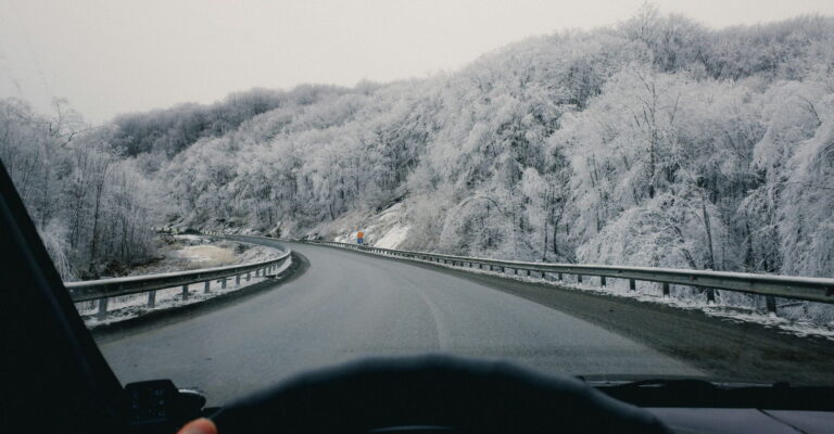 View from a car driving on a snowy, curving road lined with frosted trees. The scene is calm and serene, with a wintery, overcast sky.