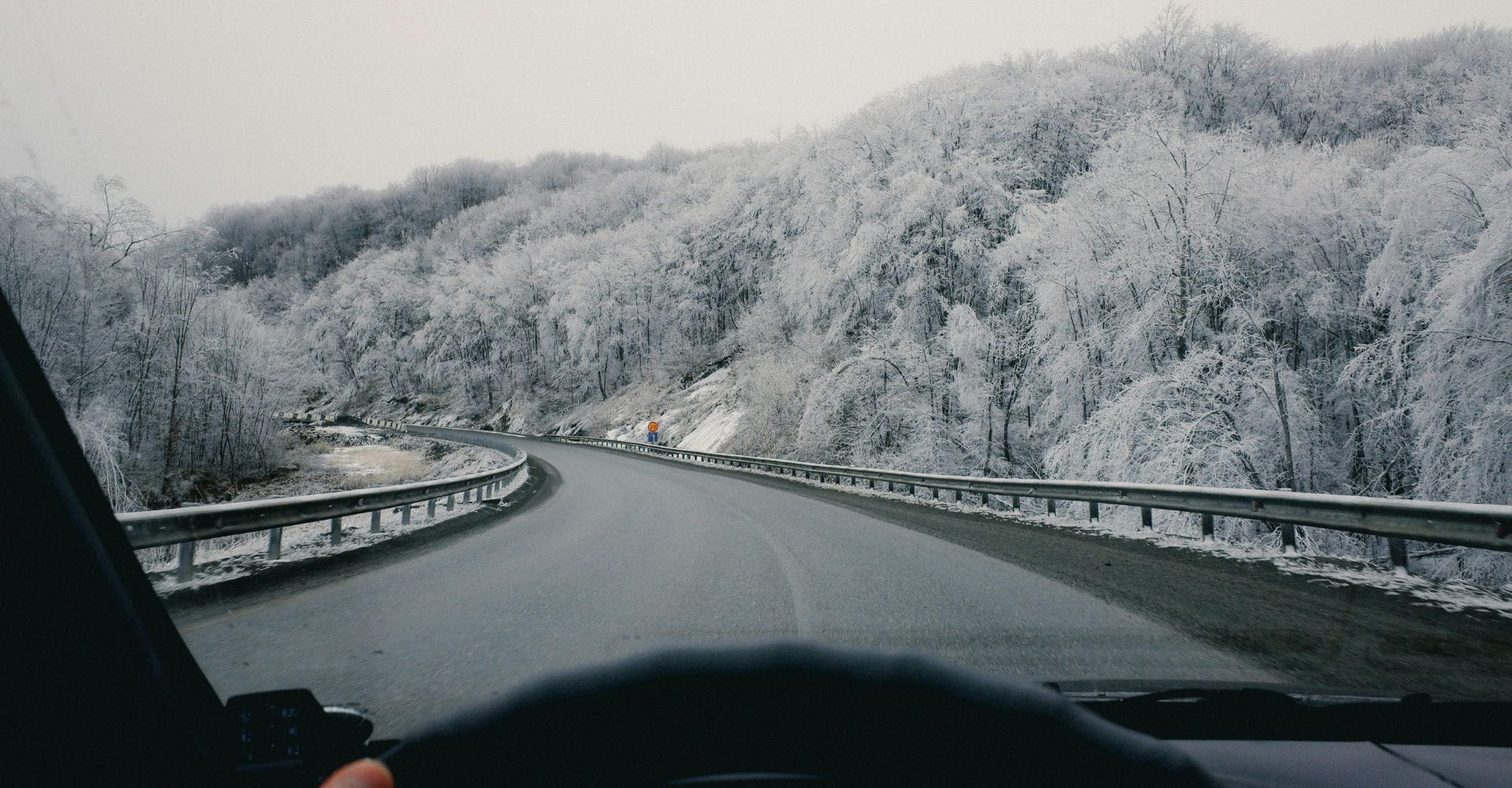 View from a car driving on a snowy, curving road lined with frosted trees. The scene is calm and serene, with a wintery, overcast sky.