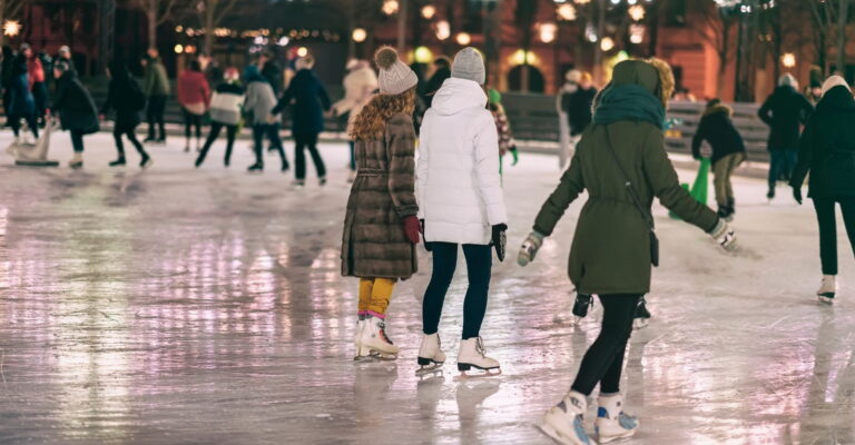 People ice skating at a rink in winter attire, including coats and hats. The scene is lively and festive, with soft evening lights in the background.