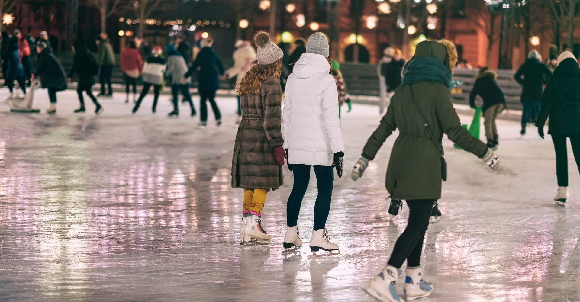 People ice skating at a rink in winter attire, including coats and hats. The scene is lively and festive, with soft evening lights in the background.