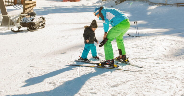 A child and an adult ski together on a sunny snow-covered slope, with ski gear and a snowmobile nearby, conveying warmth and learning.