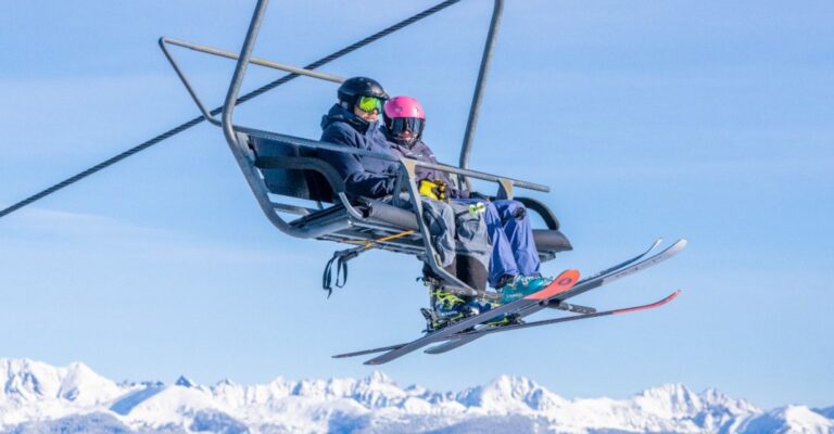 Two skiers on a chairlift with snowy mountain peaks in the background. They wear helmets and goggles, enjoying a sunny day on the slopes.