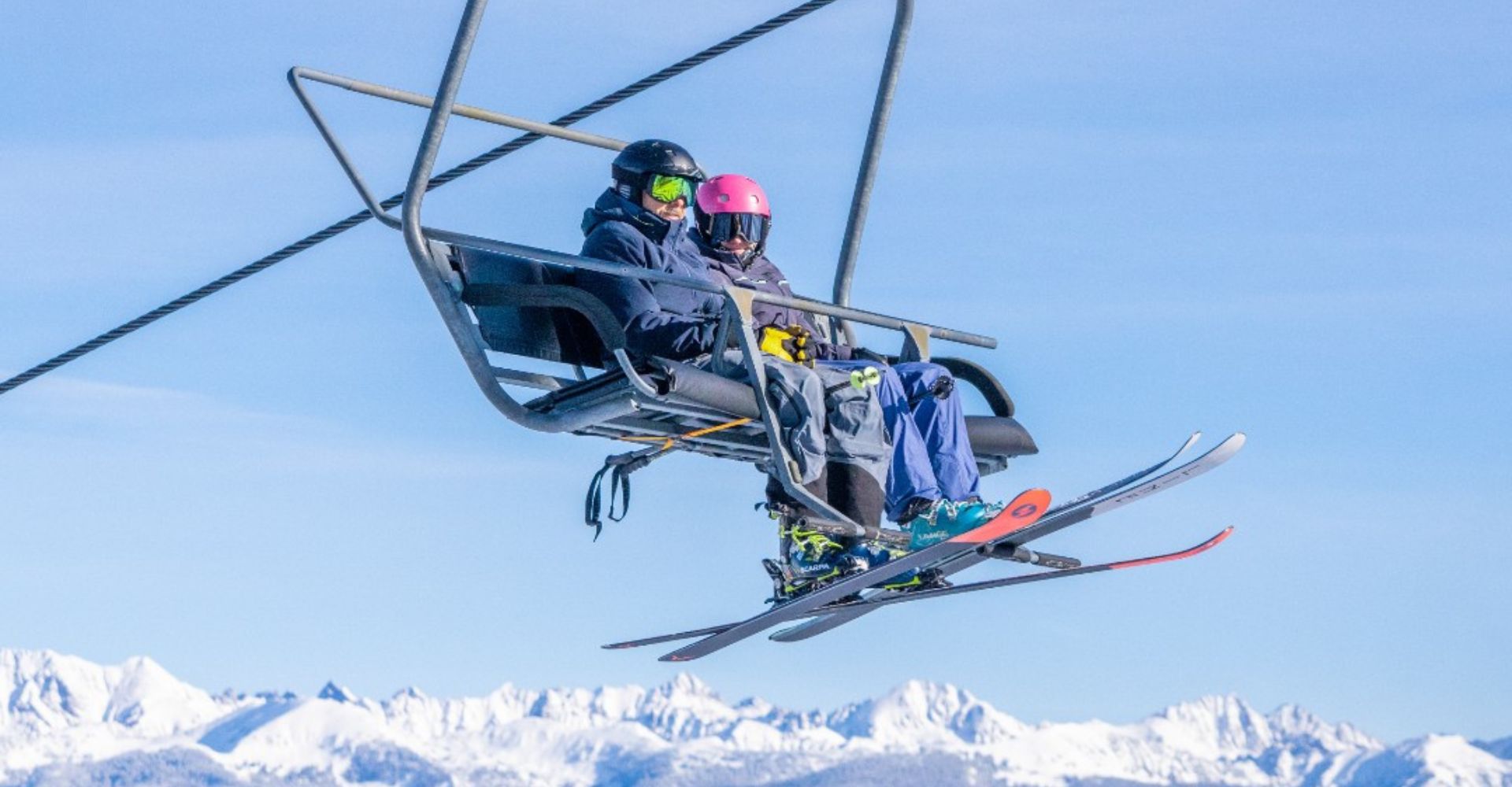 Two skiers on a chairlift with snowy mountain peaks in the background. They wear helmets and goggles, enjoying a sunny day on the slopes.