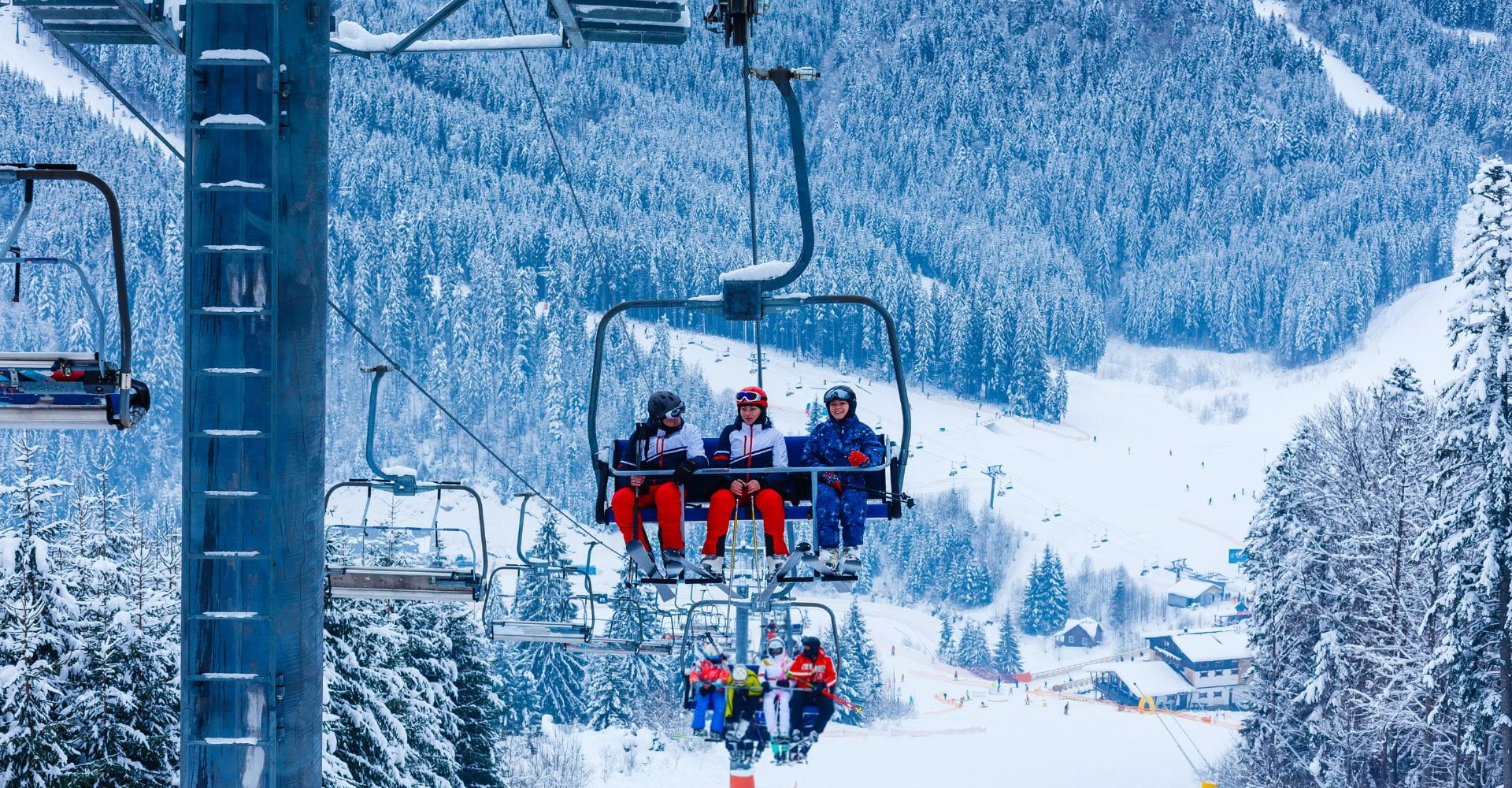 Four people in ski gear ride a snowy chairlift, surrounded by evergreen trees and snow-covered mountains, evoking a sense of adventure and excitement.
