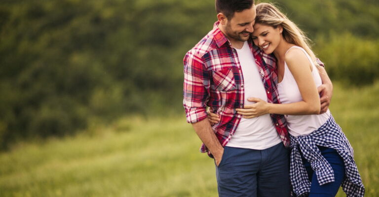A couple smiles and embraces in a lush green field. The man wears a red plaid shirt, and the woman wears a white tank top, conveying warmth and happiness.