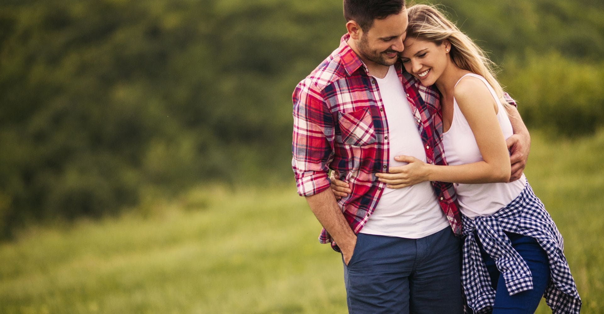 A couple smiles and embraces in a lush green field. The man wears a red plaid shirt, and the woman wears a white tank top, conveying warmth and happiness.