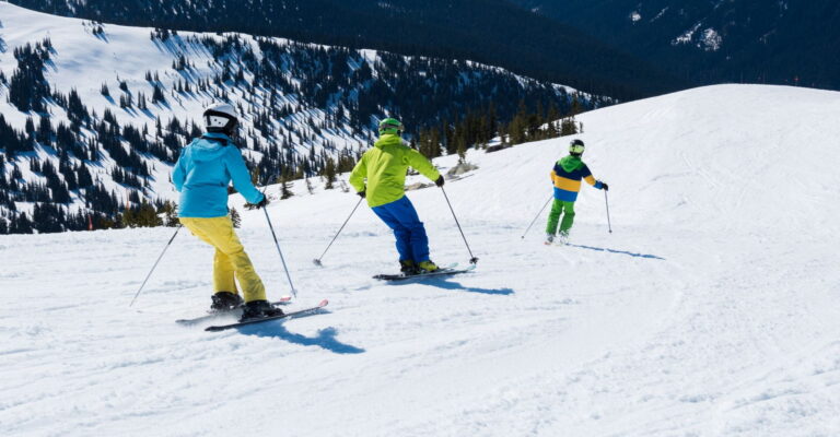 Three skiers in colorful jackets glide down a snowy mountain with evergreen trees in the background. The scene conveys a sense of adventure and fun.