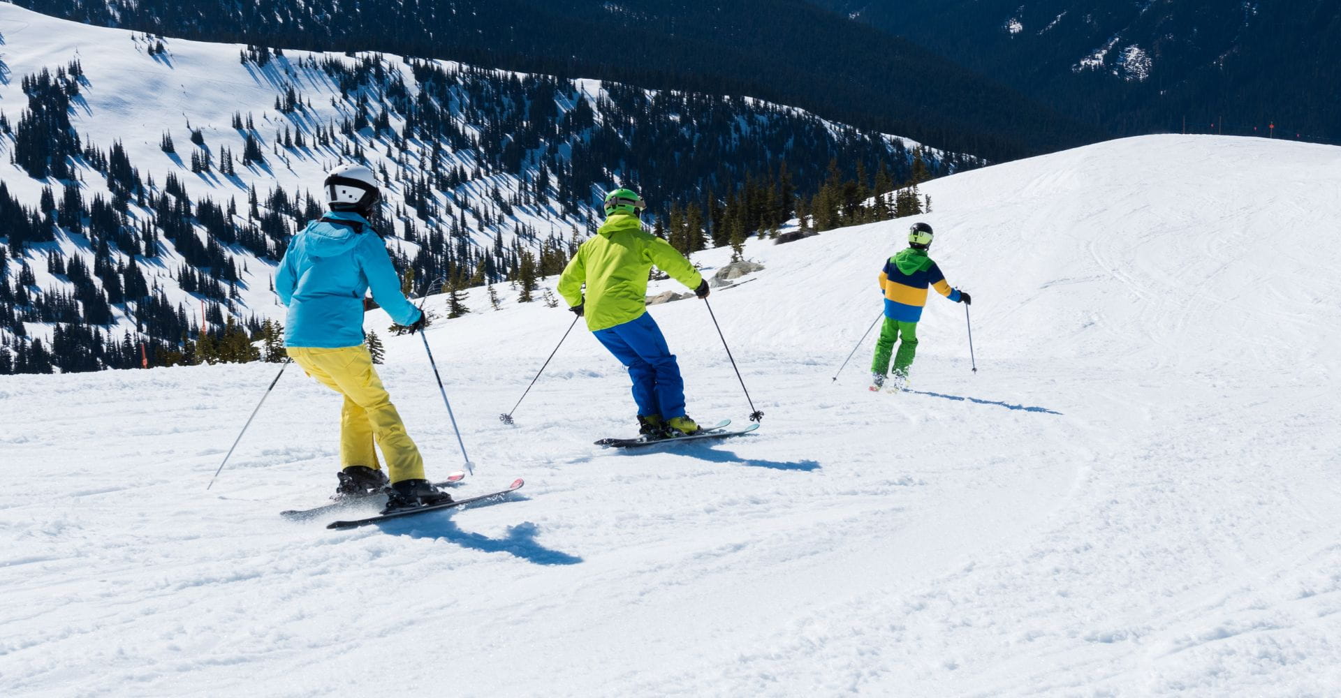Three skiers in colorful jackets glide down a snowy mountain with evergreen trees in the background. The scene conveys a sense of adventure and fun.