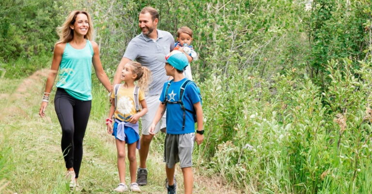 A family of four walking hand in hand along a lush green forest trail, surrounded by tall trees and sunlight filtering through leaves.